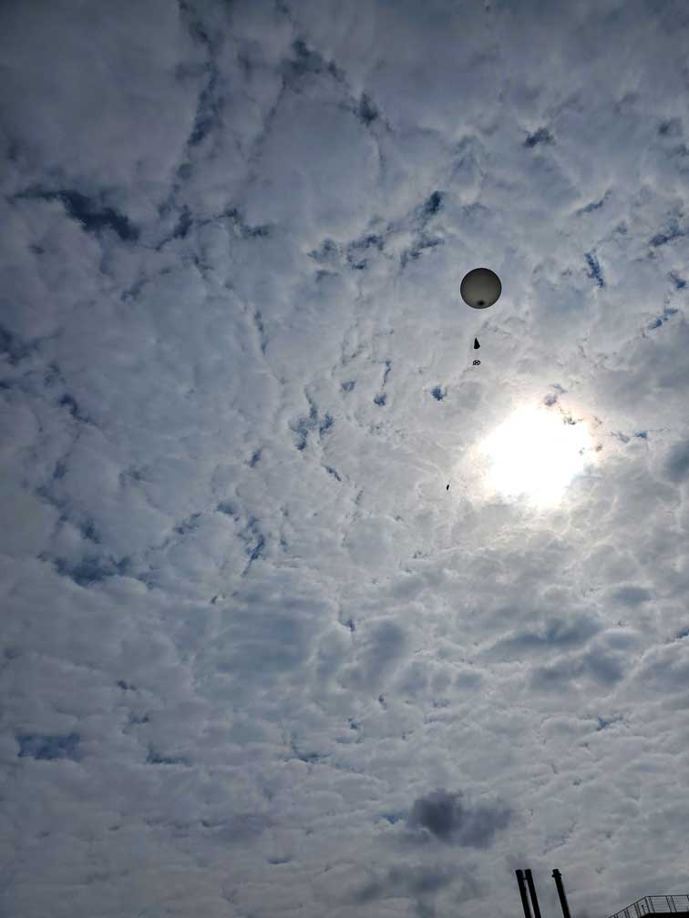 A weather balloon ascends towards the bright but cloudy sky