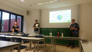 A lecture hall with black boards in the background and three people giving a presentation