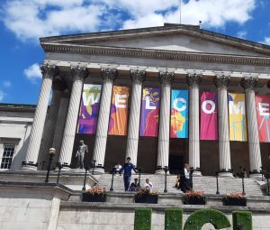 Person in front of an antic building with columns supporting a roof. Between the columns letters ar hung up forming the word welcome.
