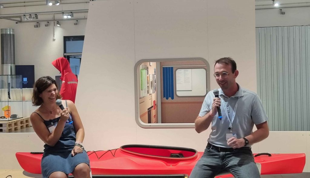 Two persons sitting in front of a kayak indoors with microphones in their hands